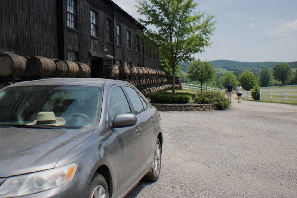 A parked car beside a barrel warehouse at a rural Kentucky distillery, with white fences and rolling hills behind it.