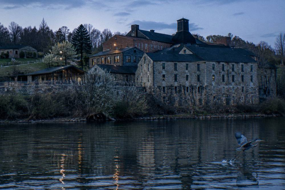 A historic brick distillery campus with stone buildings and old barrel warehouses beside a river