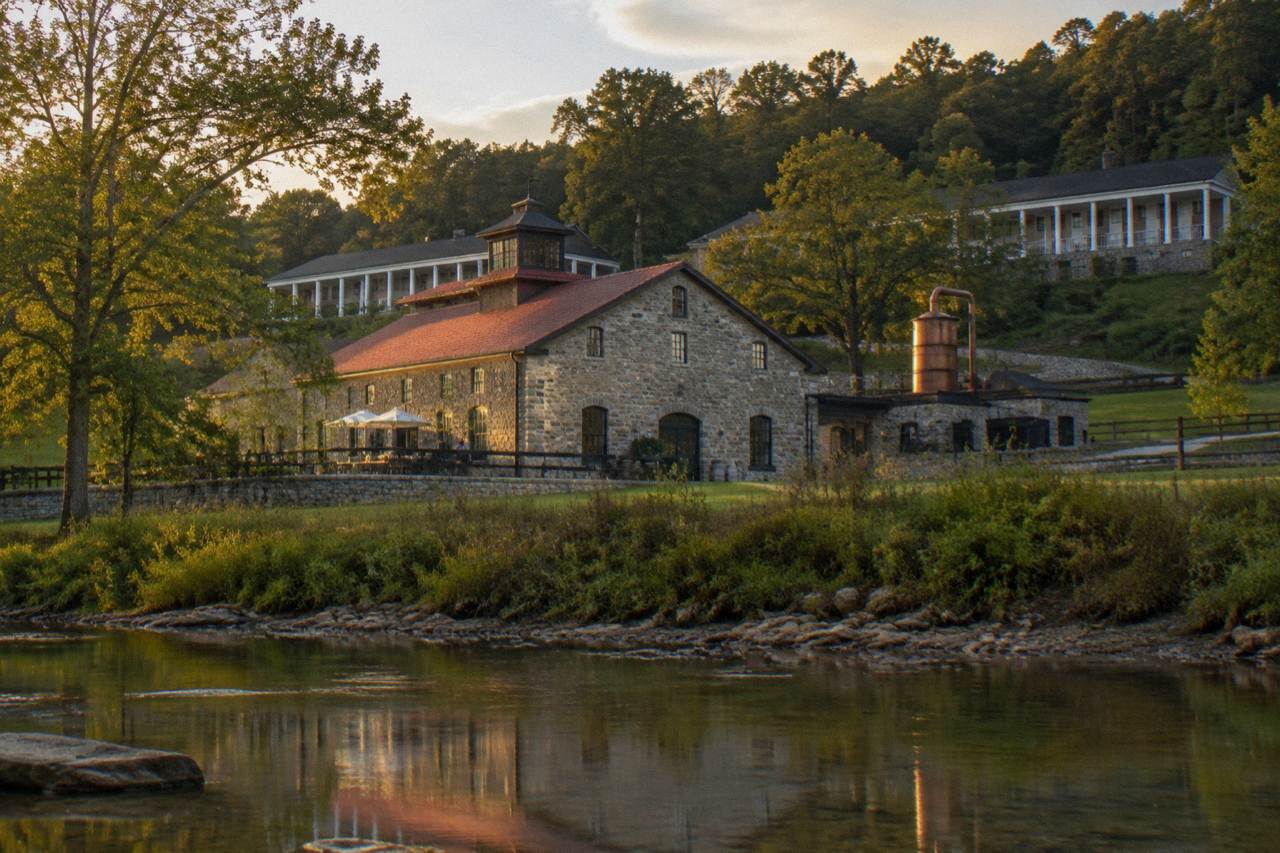 A stone distillery building beside a Kentucky creek with wooden barrel warehouses on a hillside