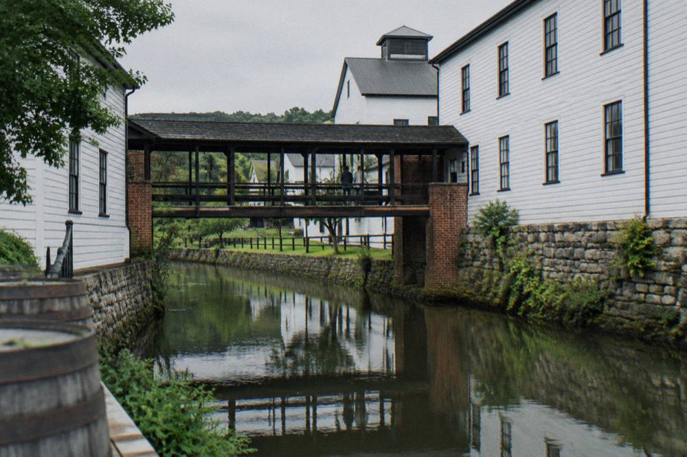 White warehouse buildings and covered brick walkways along a quiet creek at a Kentucky distillery