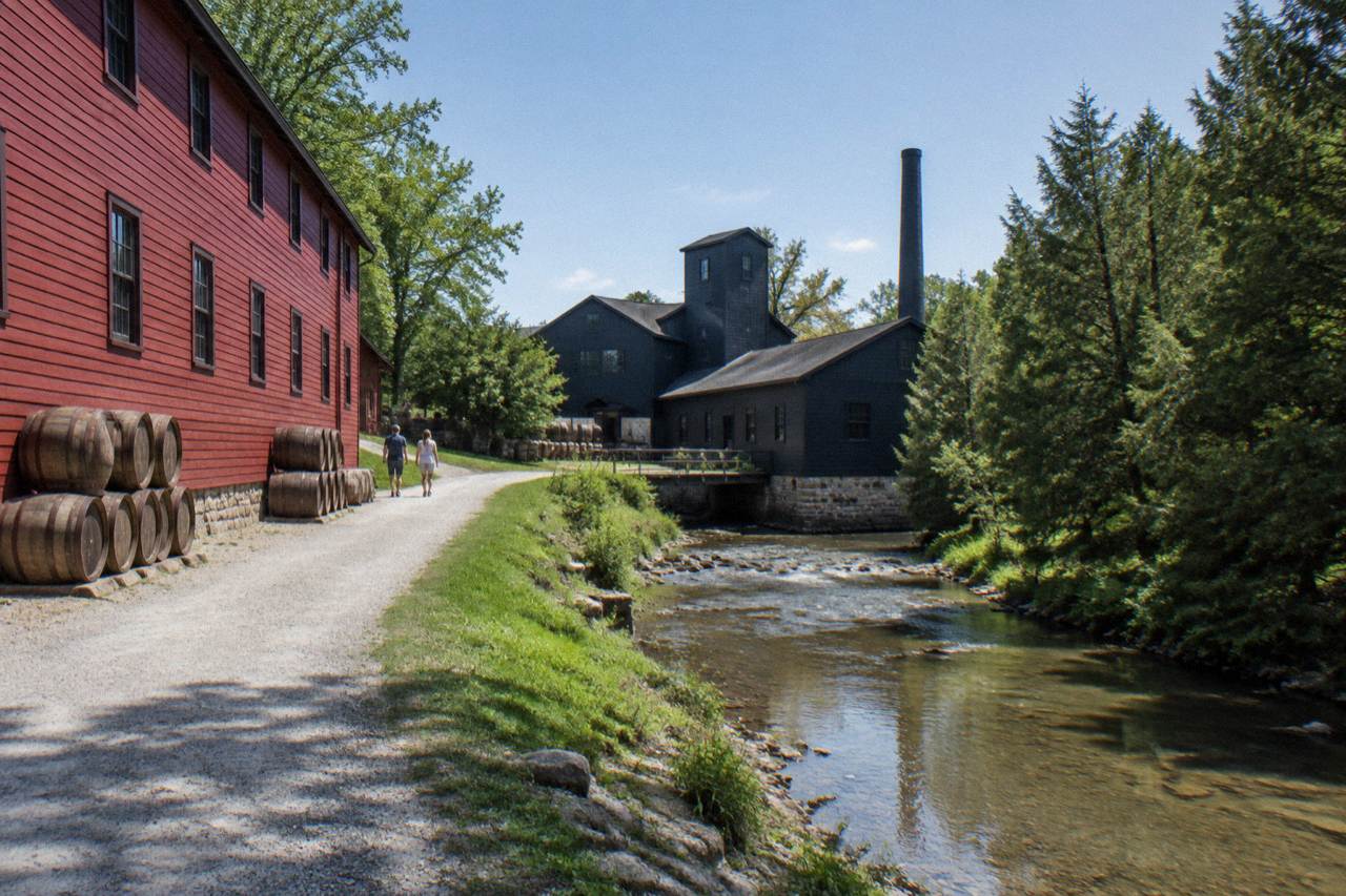 Red wooden warehouse and black distillery buildings beside a creek in the Kentucky countryside