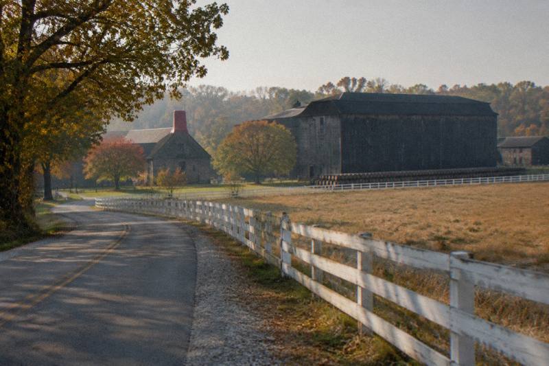 A winding Kentucky country road past a white horse farm fence and a distant barrel warehouse