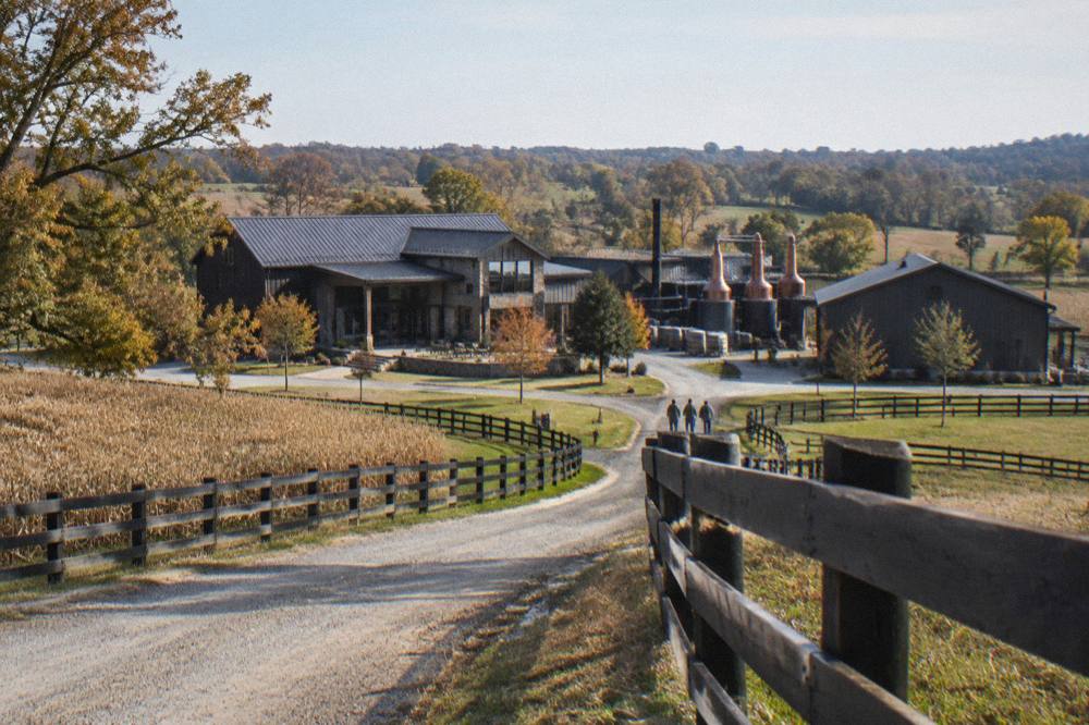 Country road curving past fenced pasture toward a distillery campus