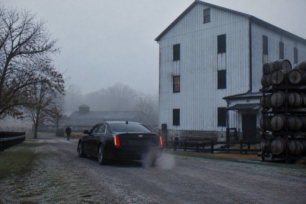 A private chauffeured vehicle waiting on a gravel drive in front of a rustic distillery rackhouse