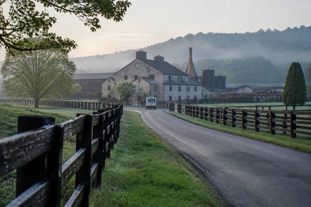 A country road winding between fenced horse farms toward a limestone distillery building