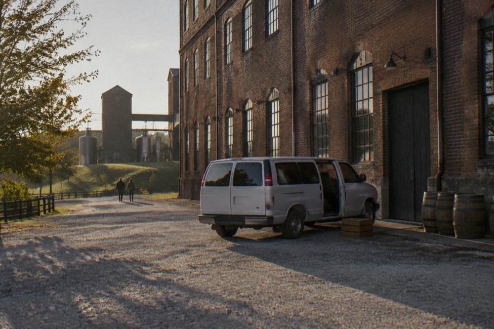 A passenger van parked on a gravel lane beside a distillery warehouse