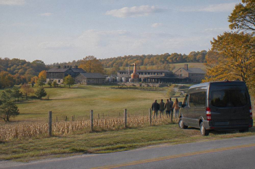 A private tour van parked on a country road with rolling hills and a distillery in the distance