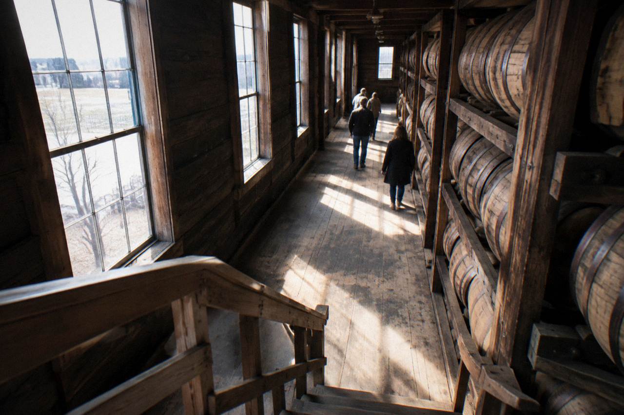 A row of bourbon barrels aging in a rickhouse with sunlight streaming through the windows
