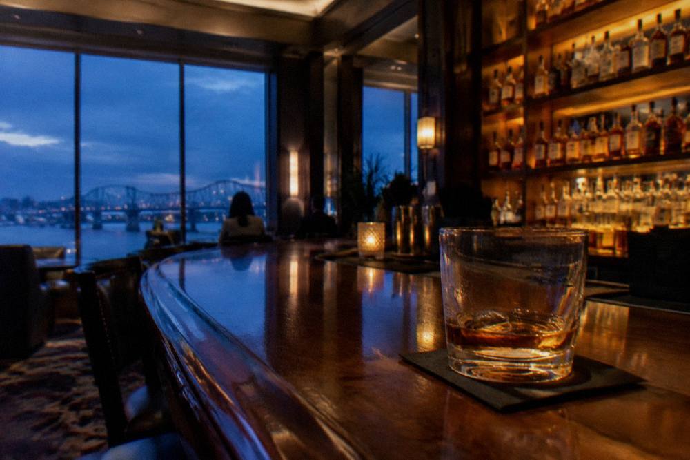 A polished wooden bar top with rows of bourbon bottles backlit on shelves