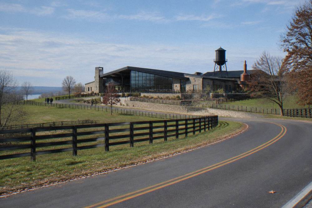Country road curving through green horse pasture toward a distillery campus