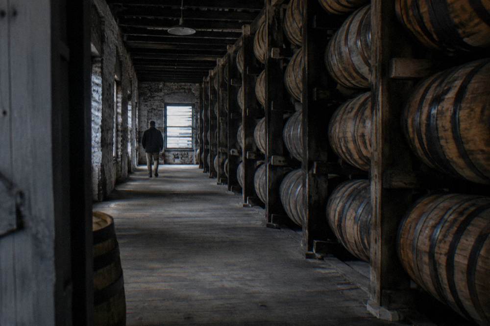 Wooden barrels stacked in rows inside a limestone maturation warehouse lit by soft daylight