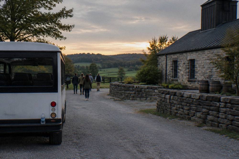 A small tour van parked on a gravel drive beside a stone distillery in green rolling hills